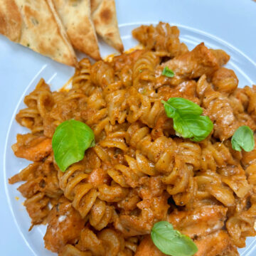 Butter chicken pasta served on a white plate, garnished with some basil leaves on top and three pita bread triangles on the side.