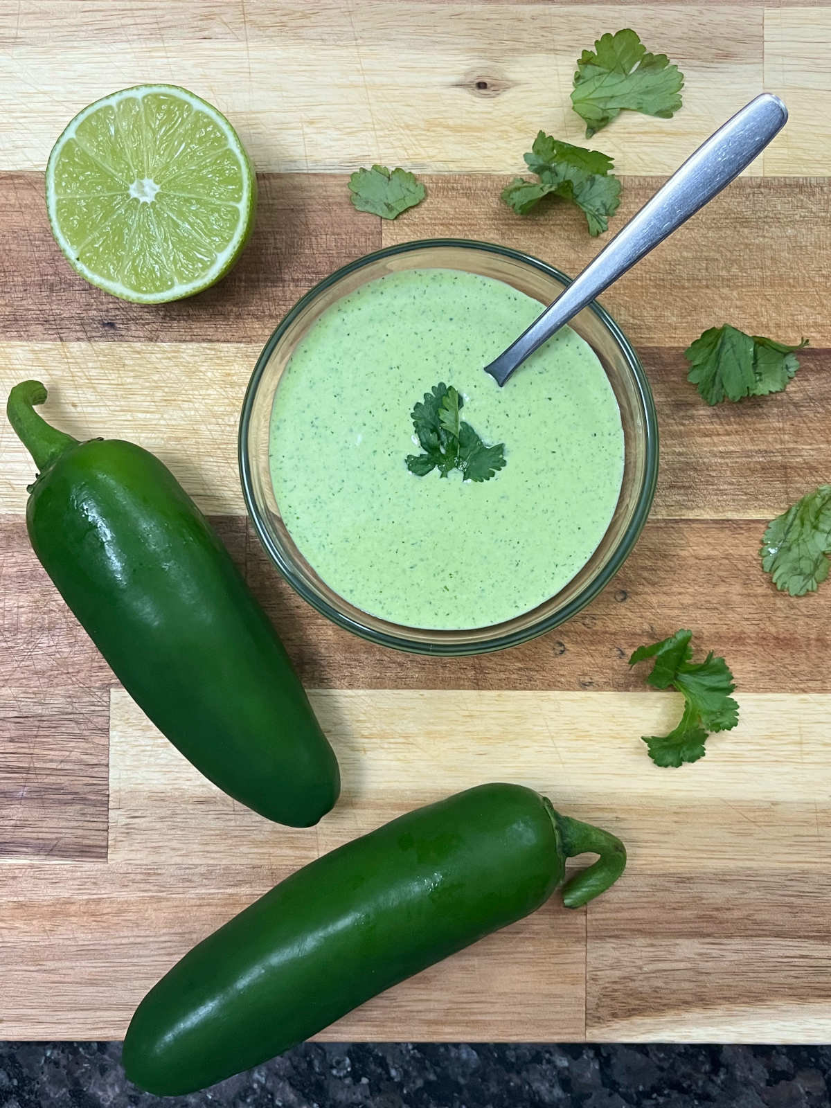 aji verde sauce in a bowl with a spoon, along with a half cut lime, two jalapeños peppers and cilantro around it