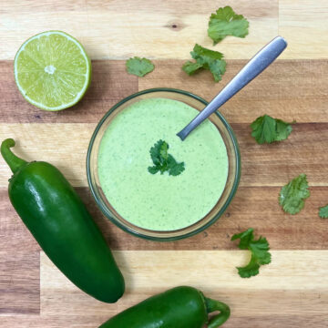 aji verde sauce in a bowl with a spoon, along with a half cut lime, two jalapeños peppers and cilantro around it