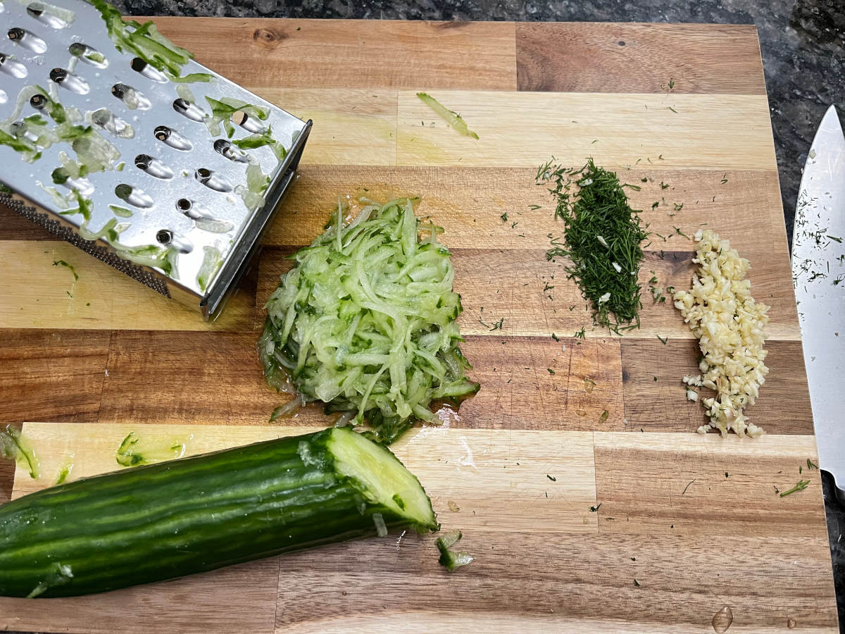 Grated cucumber with chopped garlic and dill on a cutting board. Half cut cucumber and the grater are on the cutting board as well, with a knife on the side of the cutting board.