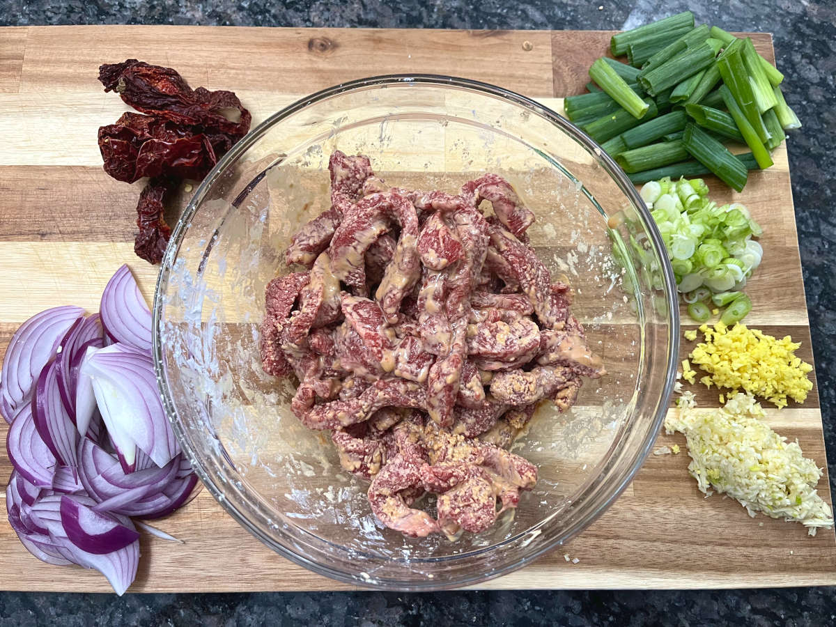 Marinated beef strips in a bowl with sliced red onions, red chilies, green onions, chopped garlic and ginger around the bowl