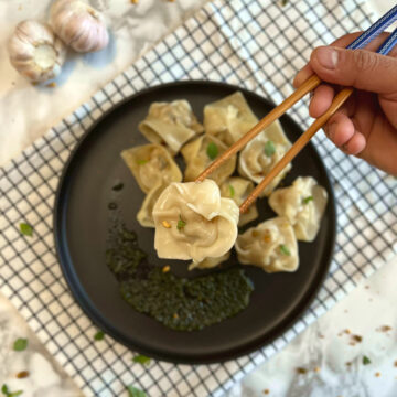 Shrimp wonton picked up by a pair of chopsticks, from a plate with wontons and pesto sauce