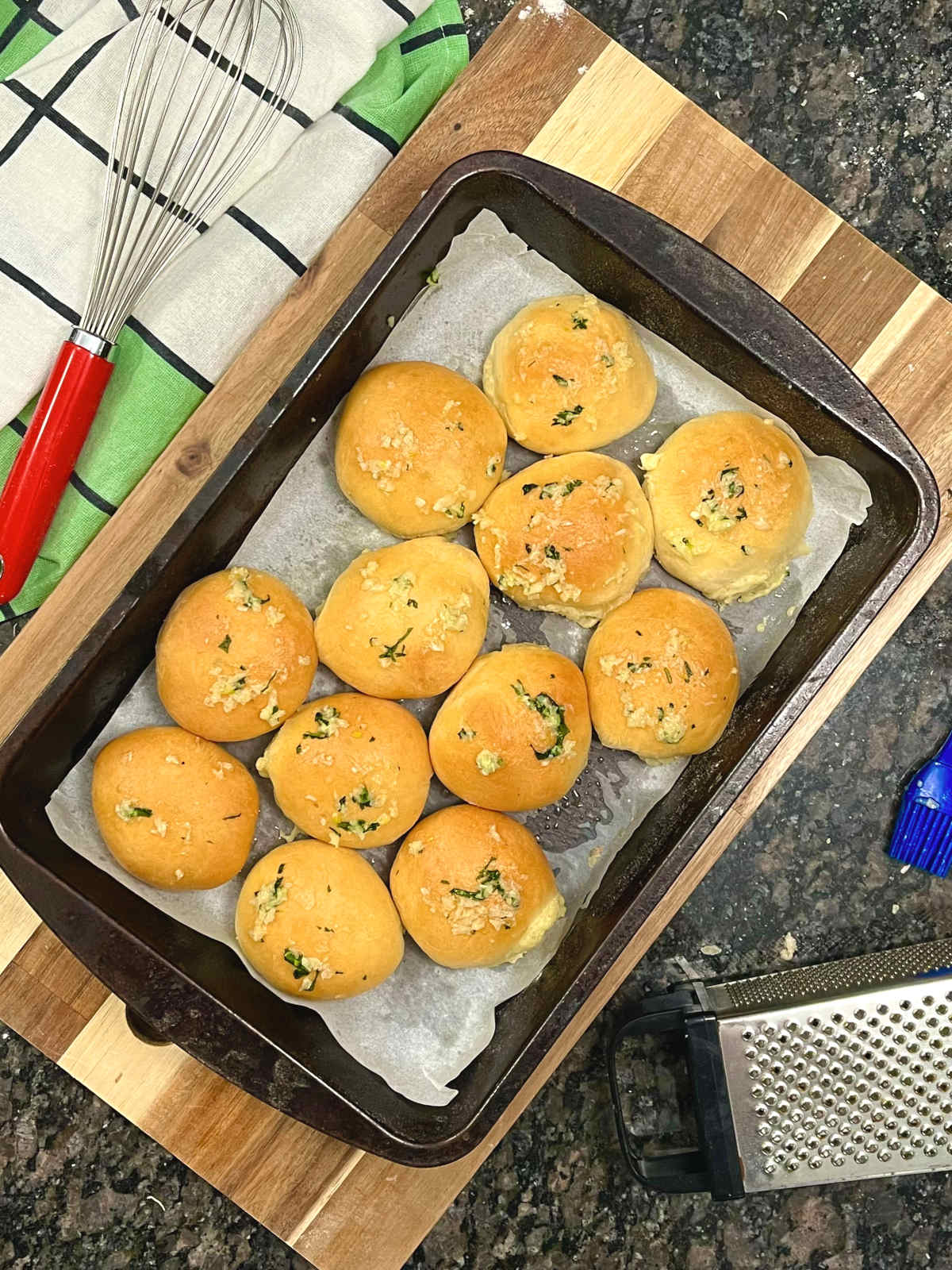 Garlic butter cheese buns in a baking tray with a whisk and grater on the side