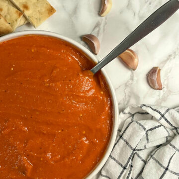 ajvar in a bowl with a spoon and some garlic and pita bread on the side